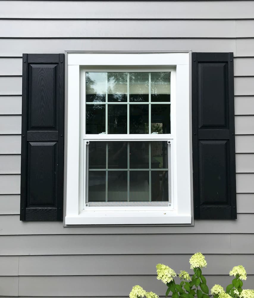 New white vinyl single-hung window with traditional grille pattern and black shutters on gray home