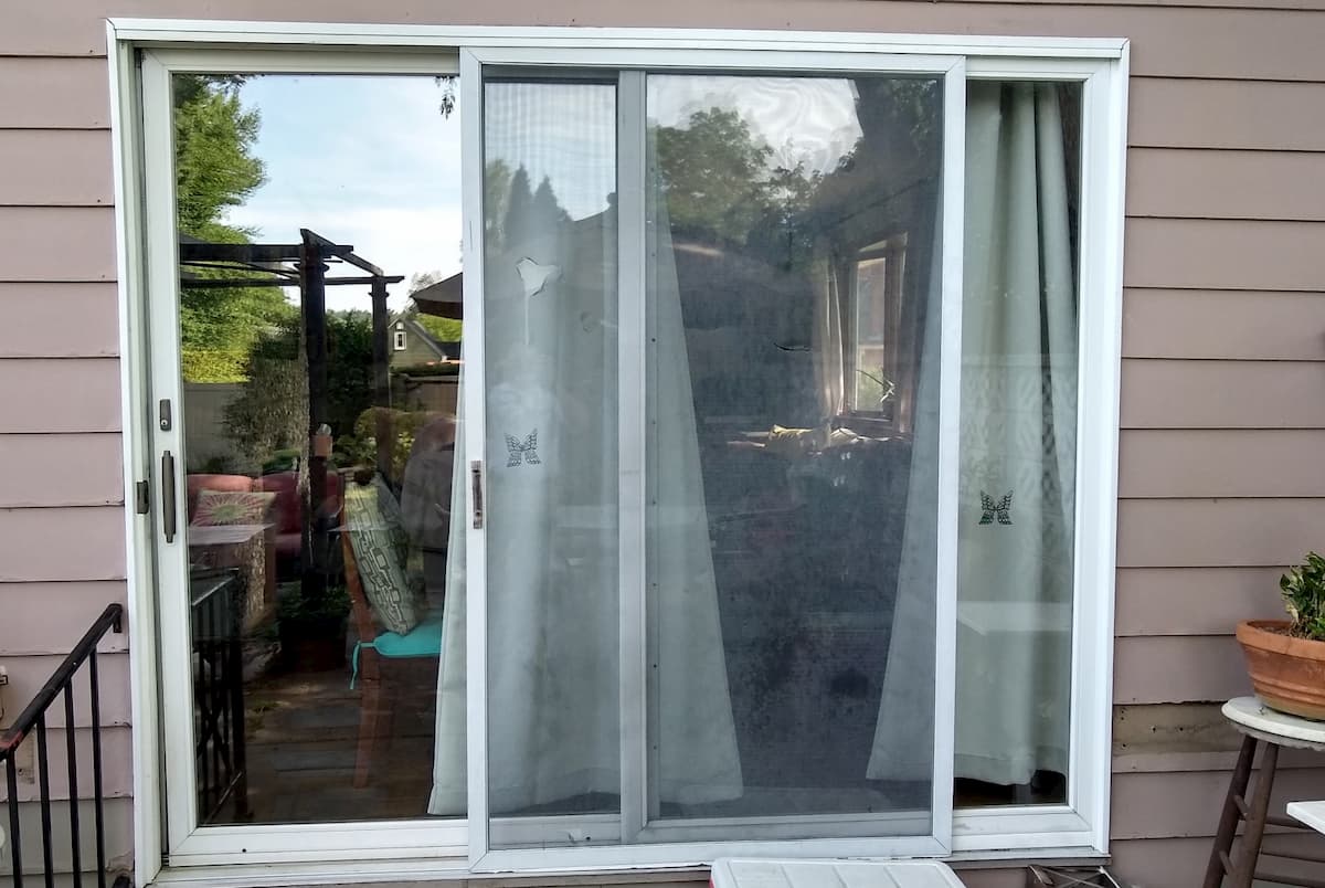 Exterior view of old white sliding patio door on a home with brown siding.