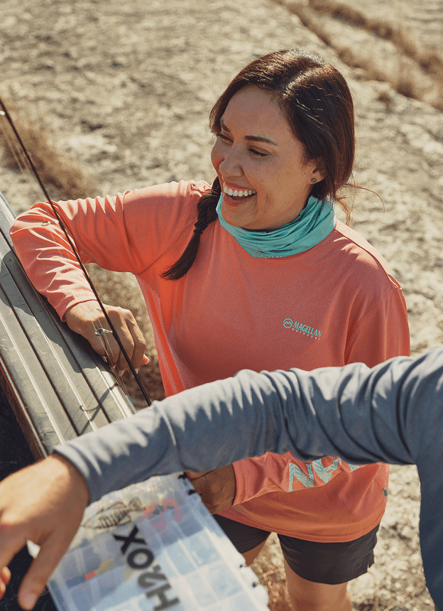 Person outdoors wearing a Magellan Outdoors long-sleeve performance shirt, leaning over a cooler near a rocky shoreline.