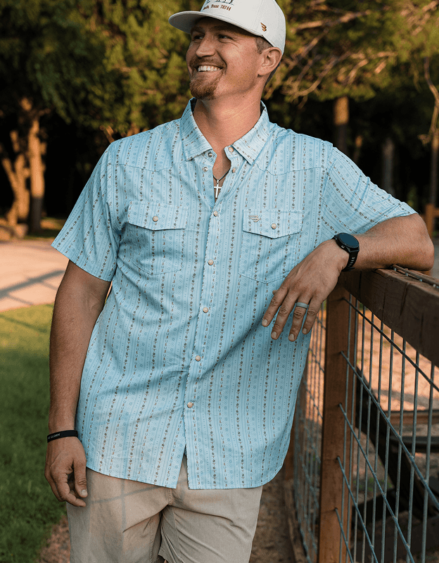 Person leaning on fence wearing a light green short sleeve western shirt outdoors.