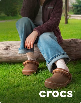 Person seated on a log outdoors wearing brown Crocs clogs, cuffed jeans, and a fleece jacket on a grassy lawn.