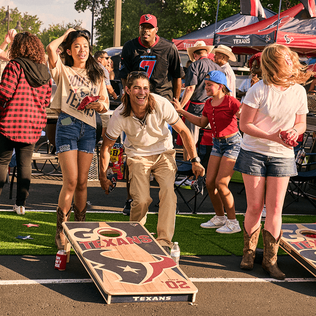 Group of fans playing cornhole at a tailgate event surrounded by team tents, games, and outdoor activities.