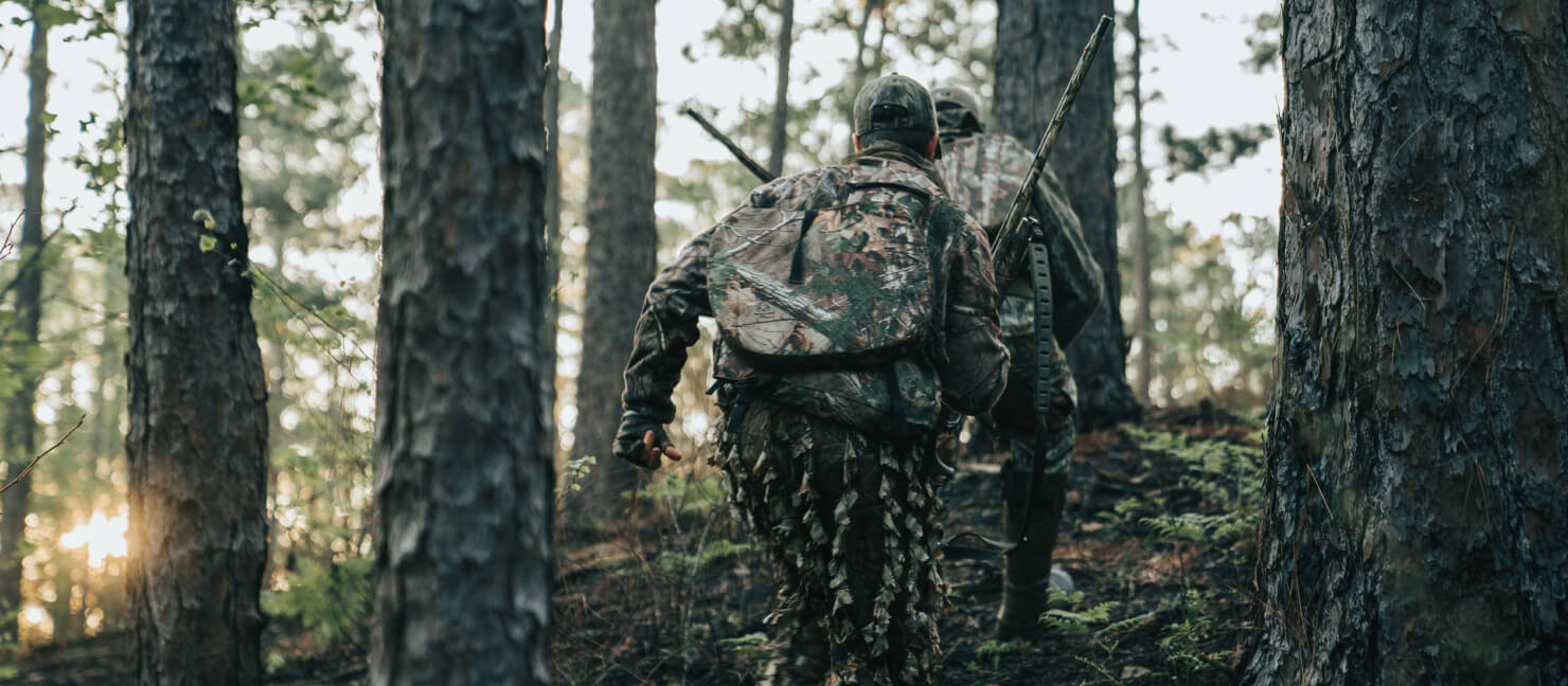 Two hunters in full camo, with shotguns and backpacks, trek up a hill in the forest.