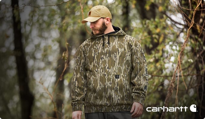 Person wearing a camouflage hooded jacket and camouflage cap standing outdoors among trees.