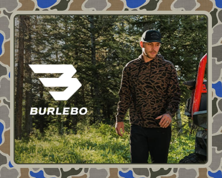 Person wearing a camouflage-pattern Burlebo pullover while standing outdoors near a pickup truck and wooded area.