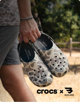 Person carrying two pairs of camouflage Crocs x Burlebo collaboration clogs outdoors.