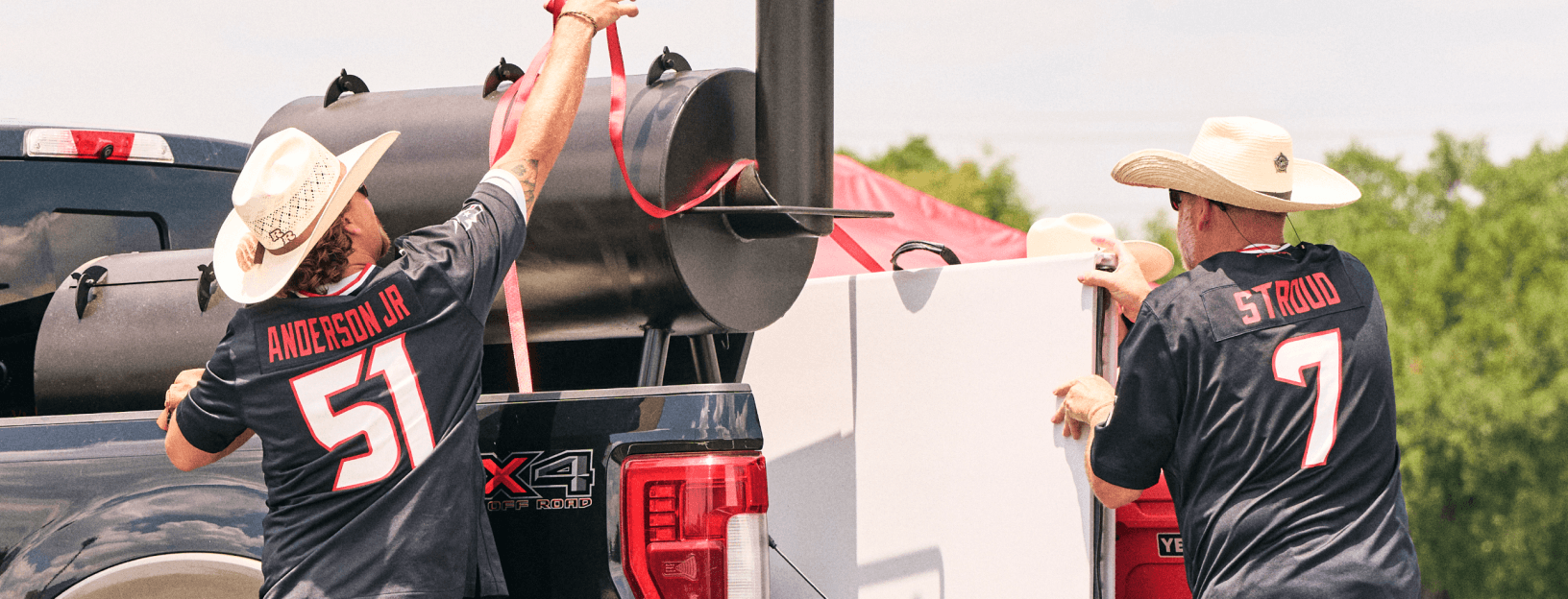 Two people wearing NFL jerseys while standing beside a large smoker grill and pickup truck outdoors, showcasing fan apparel.