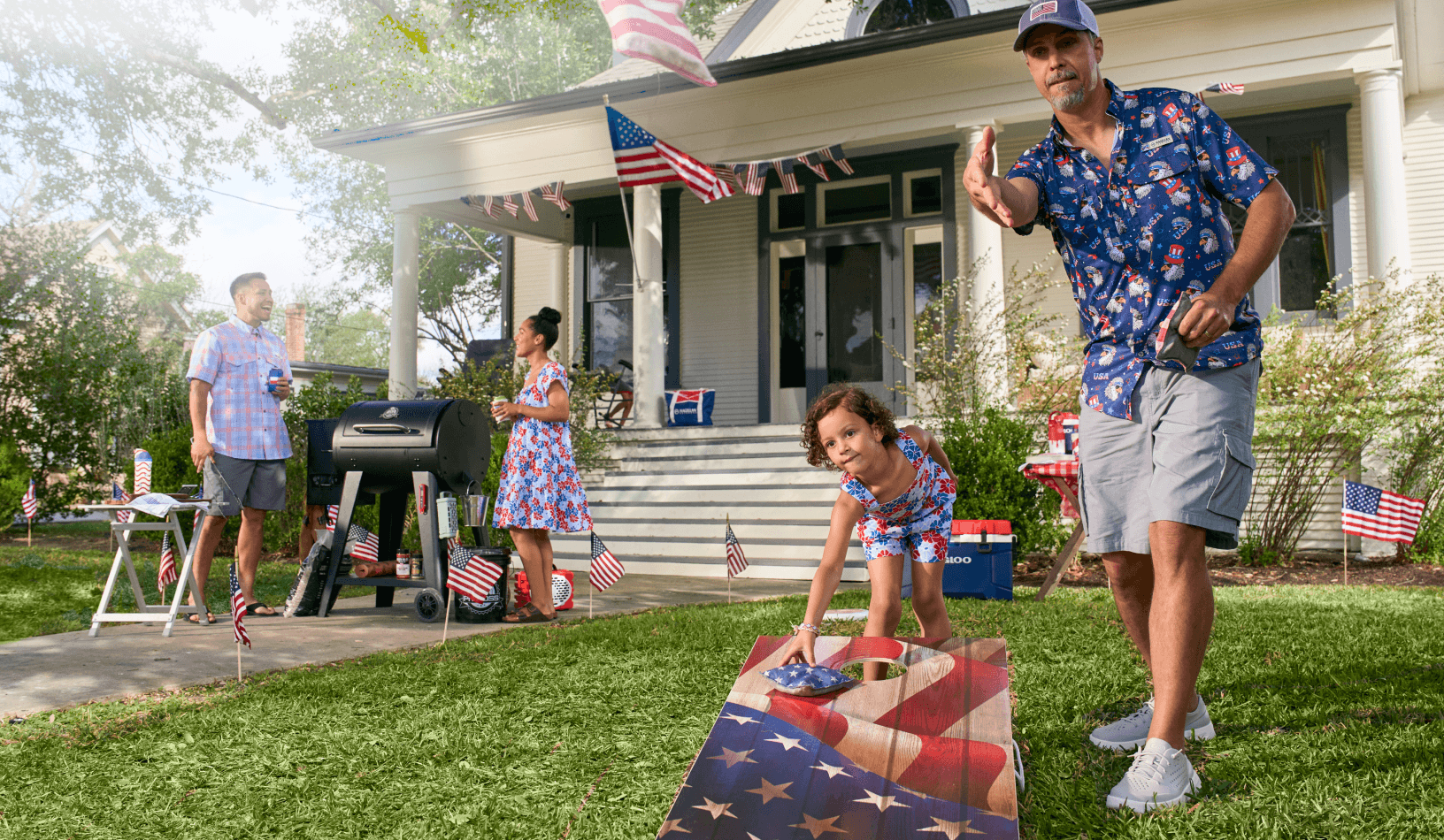 People gathered in a front yard with patriotic decorations, a grill, and a cornhole game.
