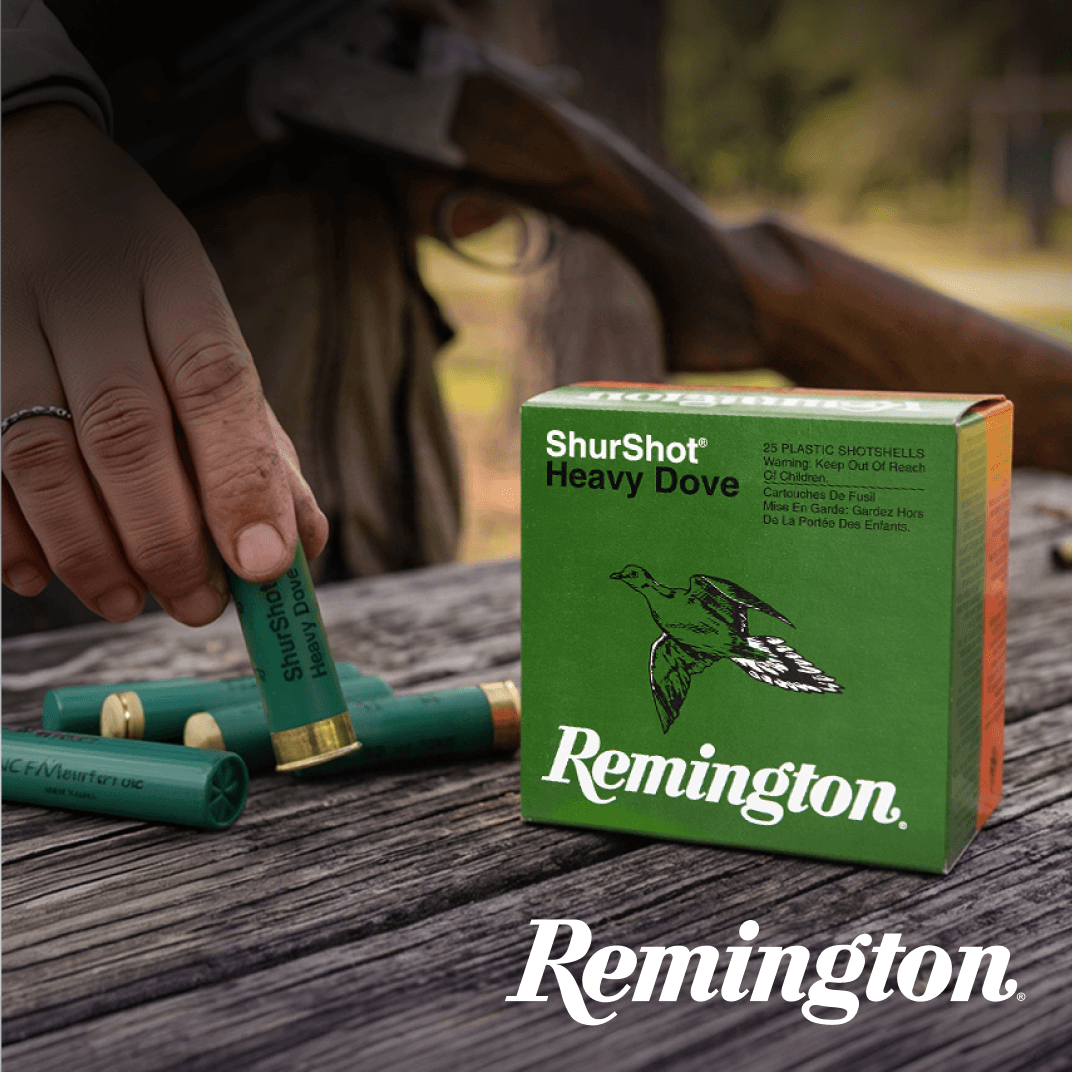 Hand loading green shotgun shells beside a box of Remington Heavy Dove shotshells on a wooden surface.