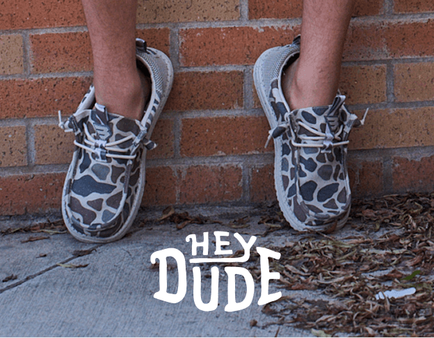 Close-up of gray HEYDUDE shoes worn with rolled jeans beside a brick wall. HEYDUDE