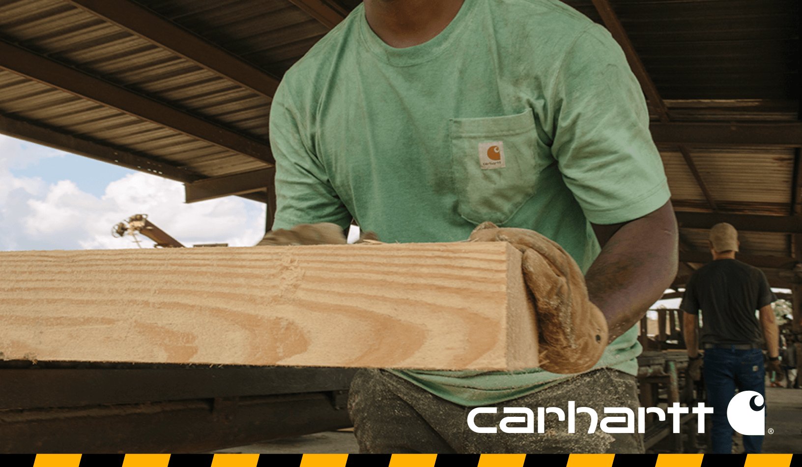 A person wearing a green Carhartt shirt and work gloves carrying a large piece of lumber in a jobsite setting, with text advertising 25% off select styles.