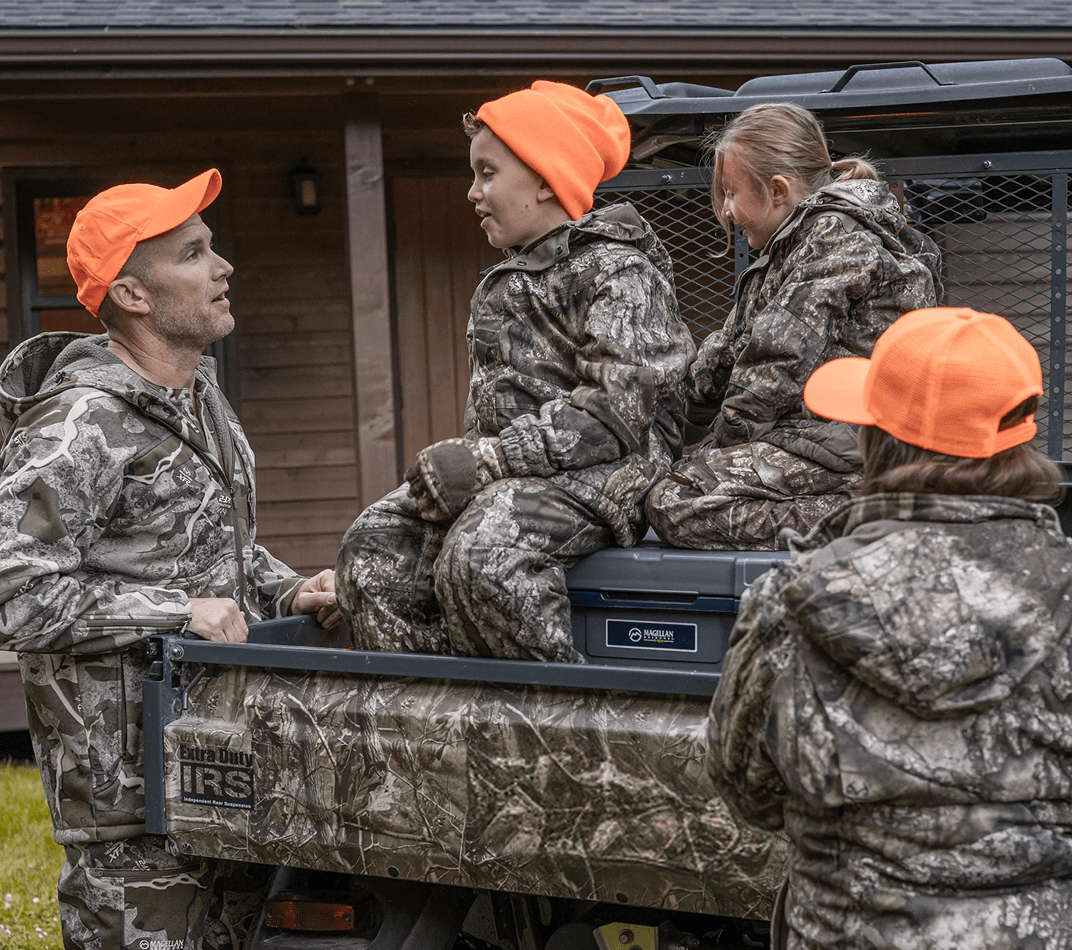 Group of people wearing Magellan Outdoors camouflage sitting in the back of a utility vehicle outdoors.