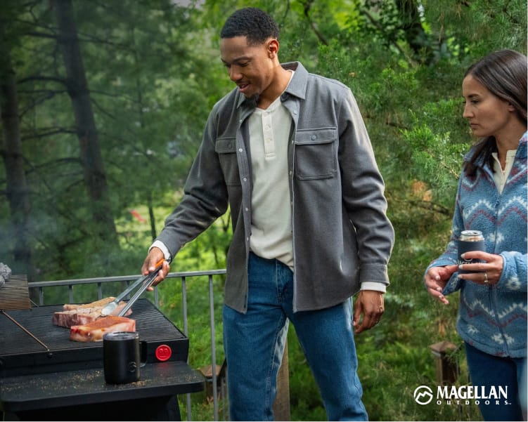 Two people standing beside an outdoor grill with food cooking, wearing Magellan Outdoors layers.