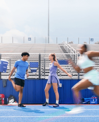 Two track athletes wearing Nike track spikes standing on a blue track while another runner speeds past, blurred from motion, with empty bleachers and sports gear in the background.