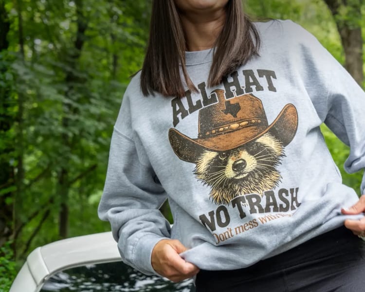 Person wearing a gray sweatshirt with a raccoon graphic and the text 'All Hat No Trash! Don't mess with Texas' while standing outdoors near a vehicle