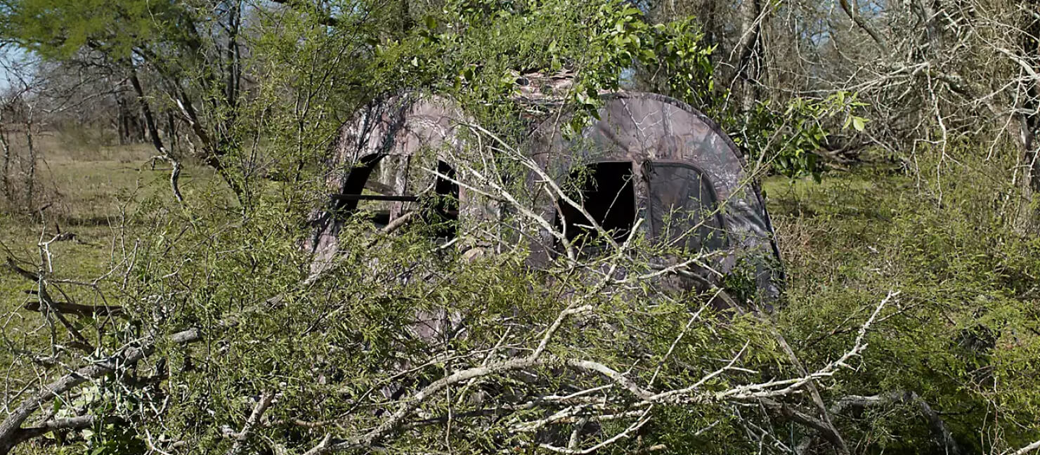 Two camo hunting blinds are hidden beneath brush in a forest.