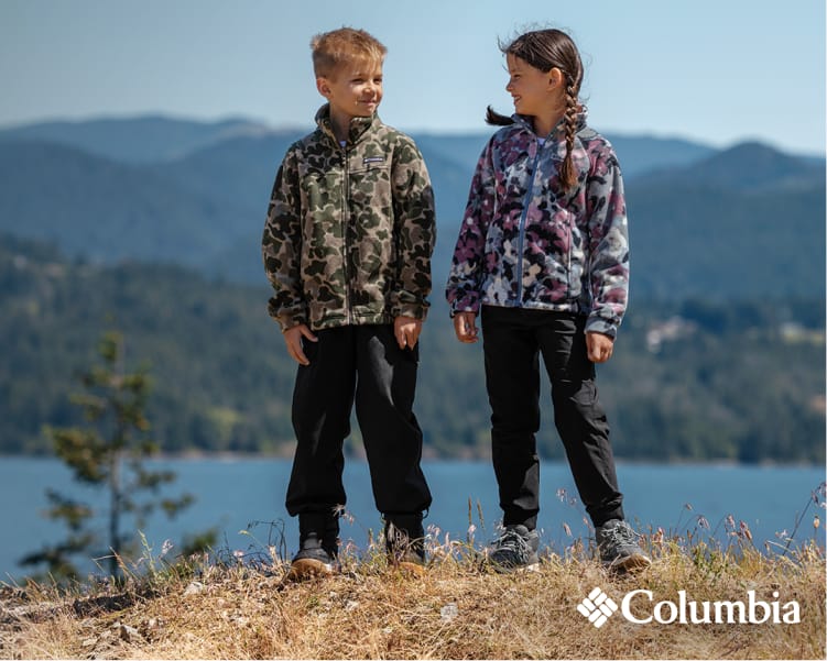 Two people wearing Columbia camouflage jackets standing outdoors near a mountain lake. Text reads: Up to 30% OFF Select Styles. Columbia logo appears in the lower-right corner.