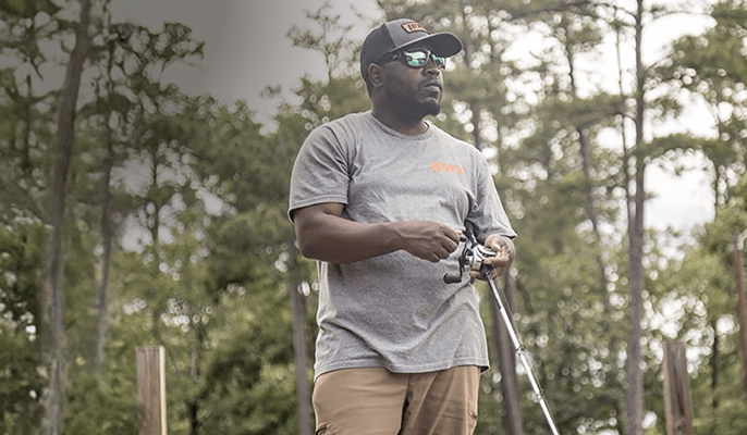 Person wearing a gray shirt and cap holding a fishing rod in an outdoor wooded setting.