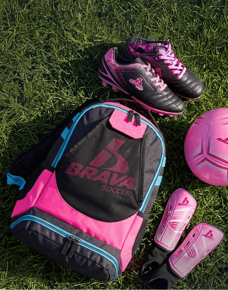 Person wearing pink soccer cleats, white knee-high socks, pink shorts, and a light pink sleeveless top jumps to strike a pink soccer ball on an outdoor turf field, with trees and a soccer goal in the background.