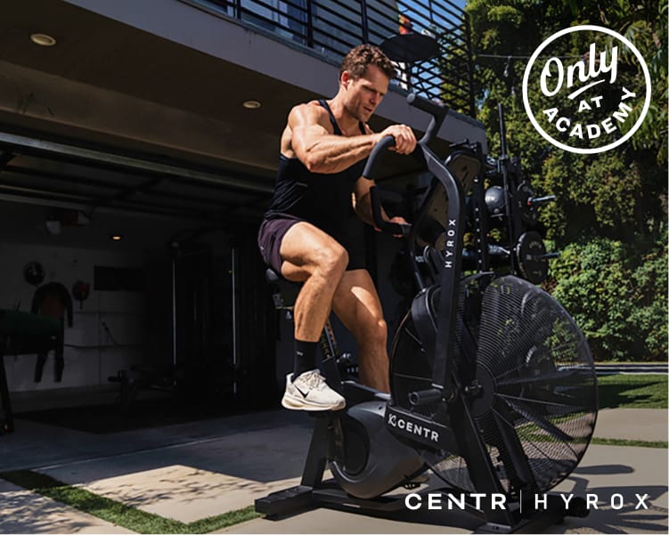 Athlete training on a CENTR HYROX air bike outside a garage gym, with overlaid text promoting new HYROX gear and an Only at Academy badge.