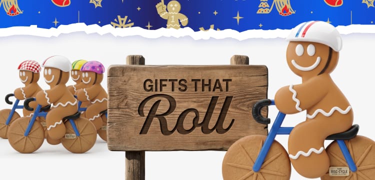 Holiday banner featuring gingerbread figures riding wooden bicycles on both sides of a wooden sign that reads Gifts That Roll. Torn-paper edges reveal blue holiday-themed borders with snowflakes and Academy Sports + Outdoors logos.