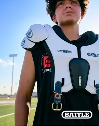 Person wearing football shoulder pads and protective gear standing on a football field. Battle Sports branding visible on the equipment.