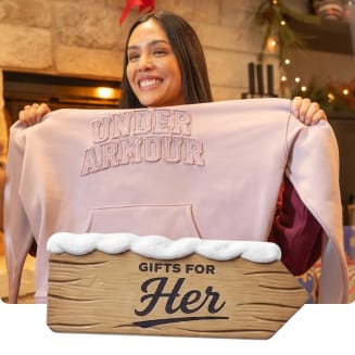 Person holding a pink Under Armour hoodie indoors near holiday decorations. A wooden holiday sign with snow reads “Gifts for Her.”