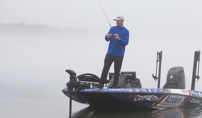 Pro angler Jacob Wheeler standing on a fishing boat holding a rod, surrounded by fog over calm water.