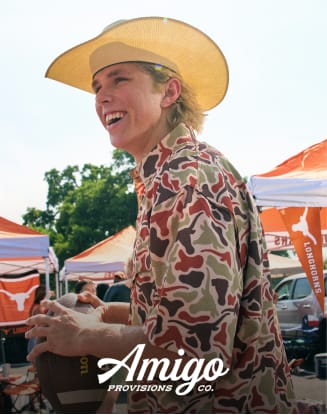 Person wearing camouflage apparel and a cowboy hat at a tailgate event. Amigo Provisions branding displayed.