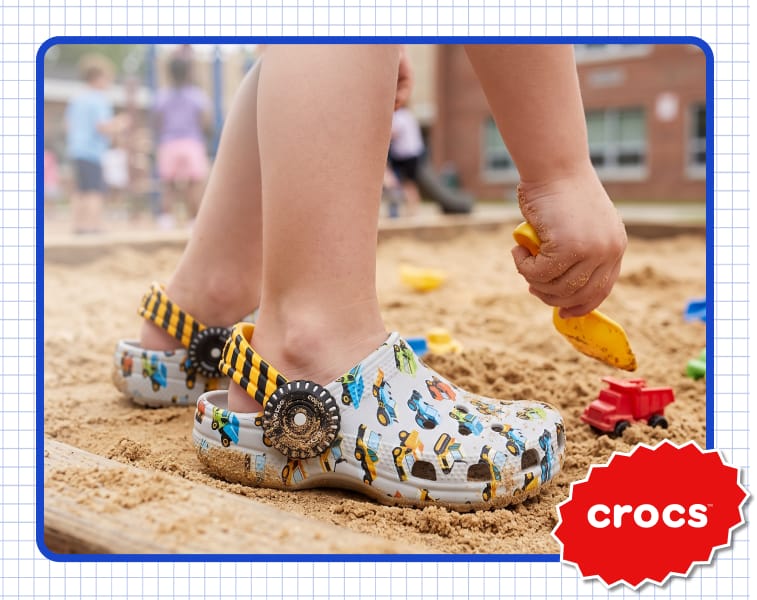 Crocs children's clogs with construction vehicle prints and black-and-yellow striped heel straps worn in a sandbox. A hand holds a yellow toy shovel above the sand, with a red toy truck nearby. Overlay