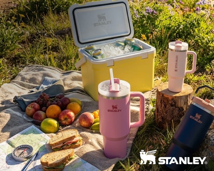 Outdoor picnic scene featuring Stanley drinkware, including tumblers and a cooler surrounded by fruit and sandwiches.