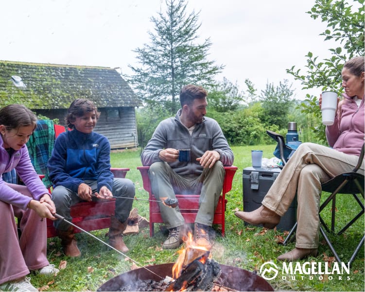 Group gathered around a campfire outside a cabin, wearing Magellan Outdoors fleece layers.