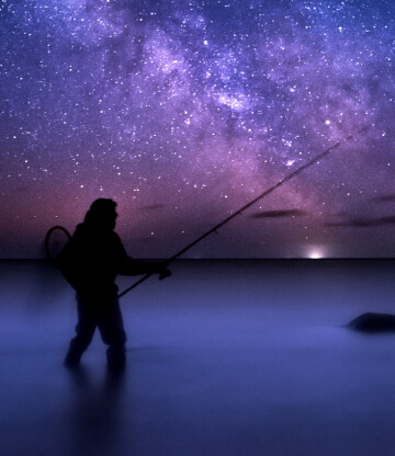 An angler fishing in saltwater under a starry night sky.