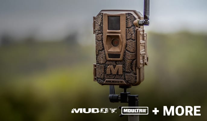 Camouflage trail camera mounted outdoors against a blurred natural background.