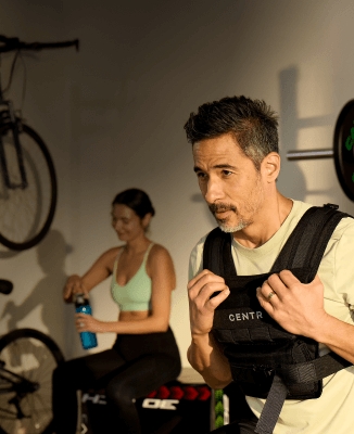 Person wearing a black weighted vest labeled 'CENTR' in their home gym, gripping its straps, with weightlifting equipment and another individual holding a blue water bottle in the background.
