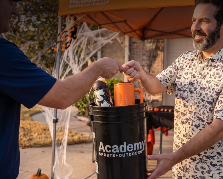 Customer receiving a Halloween tumbler from an outdoor setup beside an Academy-branded bucket during a camping or tailgating activity.