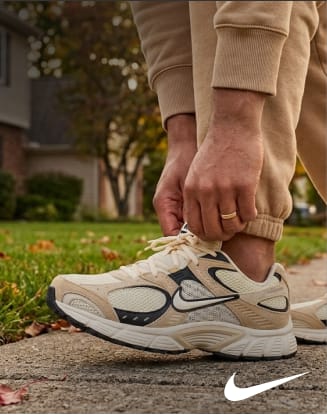 Close-up of a person tying the laces of a beige, gray, and black Nike athletic shoe while standing on a sidewalk next to a grassy lawn. The white Nike Swoosh logo appears in the lower-right corner.