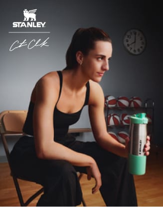 Caitlin Clark seated indoors holding her collaboration's Stanley tumbler.