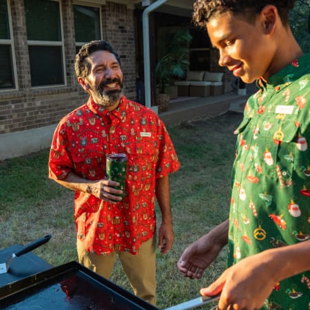 Person wearing a red Magellan Outdoors holiday button-down shirt with festive prints stands beside an outdoor griddle.