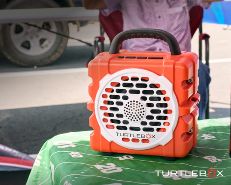 Portable Turtlebox speaker sitting on a tailgating table decorated with a football field-themed table cover beside a vehicle.