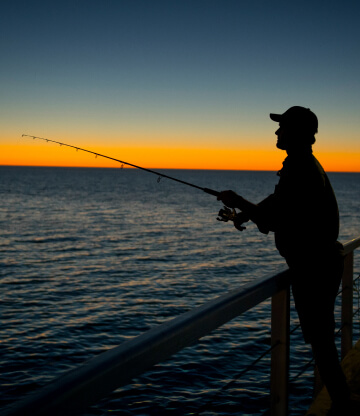 A male angler fishing on a pier at sunset.