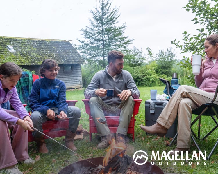 Family gathered around a campfire at a campsite with outdoor chairs and trees in the background. Magellan Outdoors logo appears in the lower-right corner.