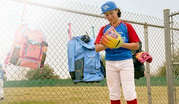 Baseball player in a red and blue uniform holding a colorful glove while standing in front of a chain-link fence with equipment bags hanging behind.