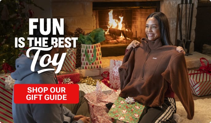 Holiday gift-opening scene in a living room with a lit fireplace and wrapped presents. One person sits on the floor holding up a brown pullover while another person sits nearby among opened gifts and wrapping paper. Promotional text reads: FUN IS THE BEST Toy. Red button reads: SHOP OUR GIFT GU