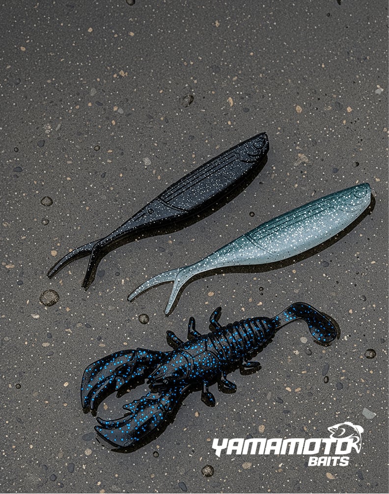 Three Yamamoto soft‑plastic fishing baits on wet pavement, including two minnow‑style lures and one crawfish‑style lure.