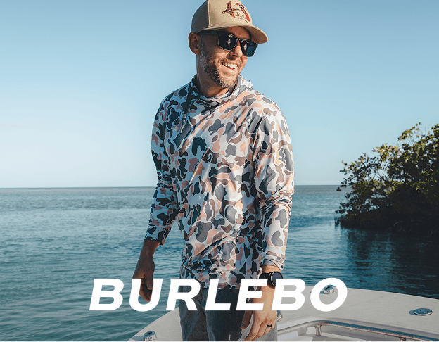 Person wearing a BURLEBO camouflage-pattern shirt and hat standing outdoors near water and trees.