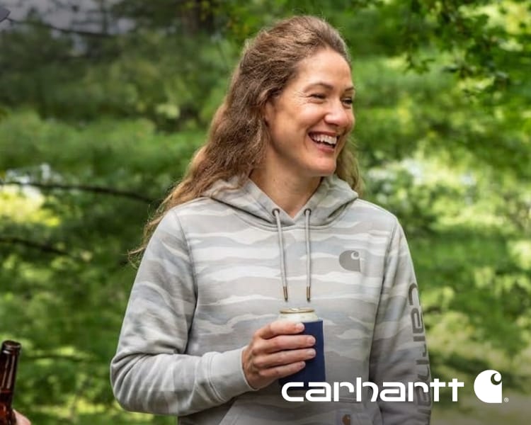 Person wearing a light gray Carhartt hoodie outdoors while holding a beverage. Carhartt logo appears in the lower-right corner.