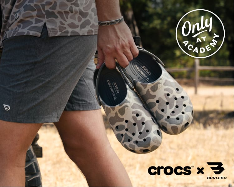 Person wearing a camouflage long-sleeve shirt and gray shorts holds a pair of Crocs Classic Clogs in a tan and gray Classic Deer Camo pattern outdoors. White overlay text reads, “Two Classics Classic Deer Camo + Classic Clog.” A circular badge reads, “Only at Academy.” Crocs x Burlebo logo appears in the bottom right corner.