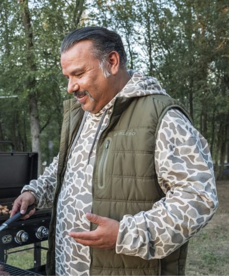 Person grilling outdoors near a campsite, wearing a camo hoodie and olive quilted vest