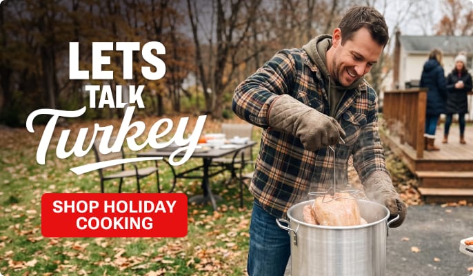 Person lowering a turkey into a large outdoor frying pot in a backyard with autumn trees and family gathering in the background. Promotional text reads: LETS TALK Turkey. Red button reads: SHOP TURKEY FRYING.